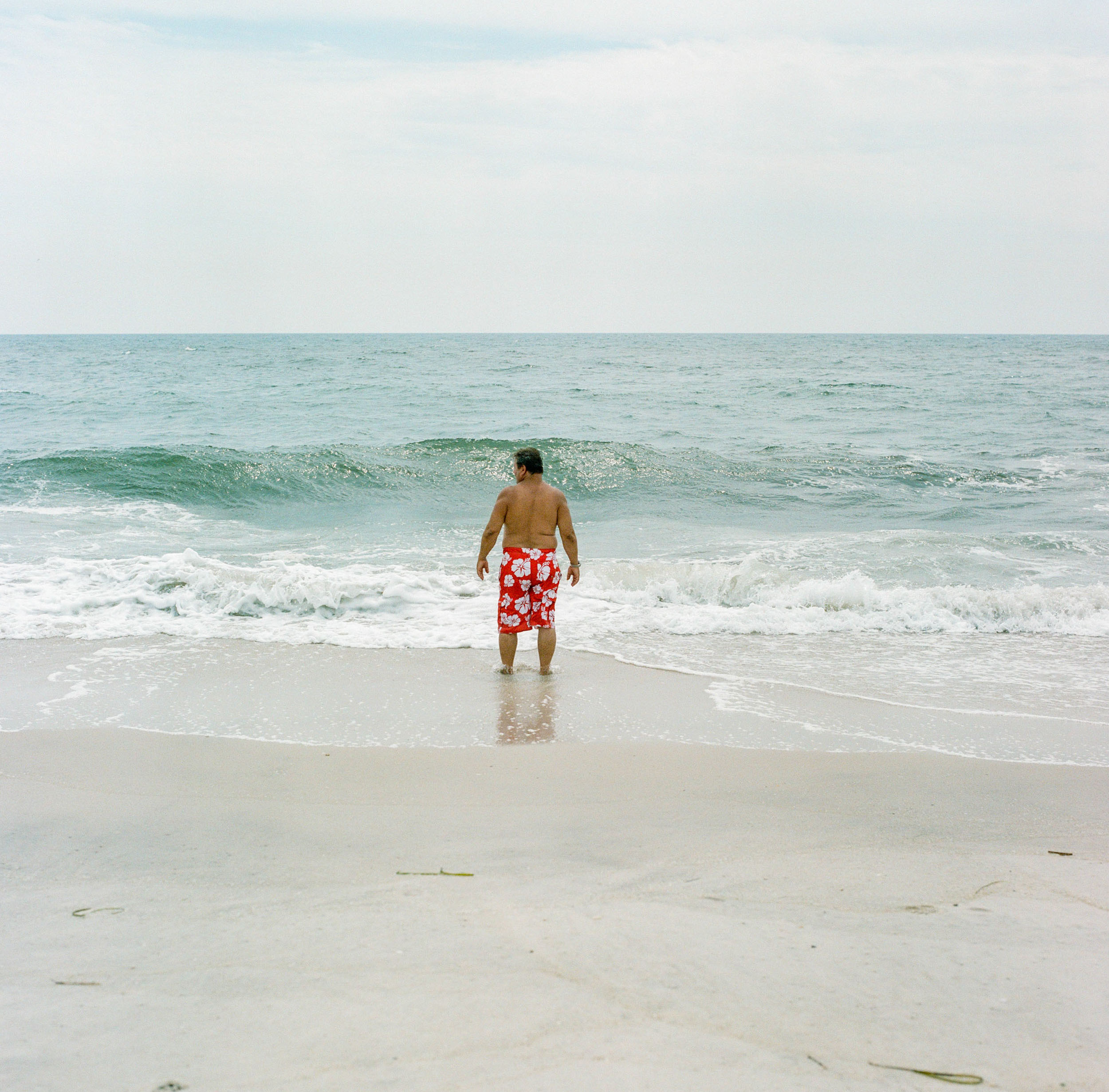 A MAN IN SWIM SHORTS STANDS IN FRONT OF THE OCEAN IN FIRE-ISLAND NEW-YORK_1